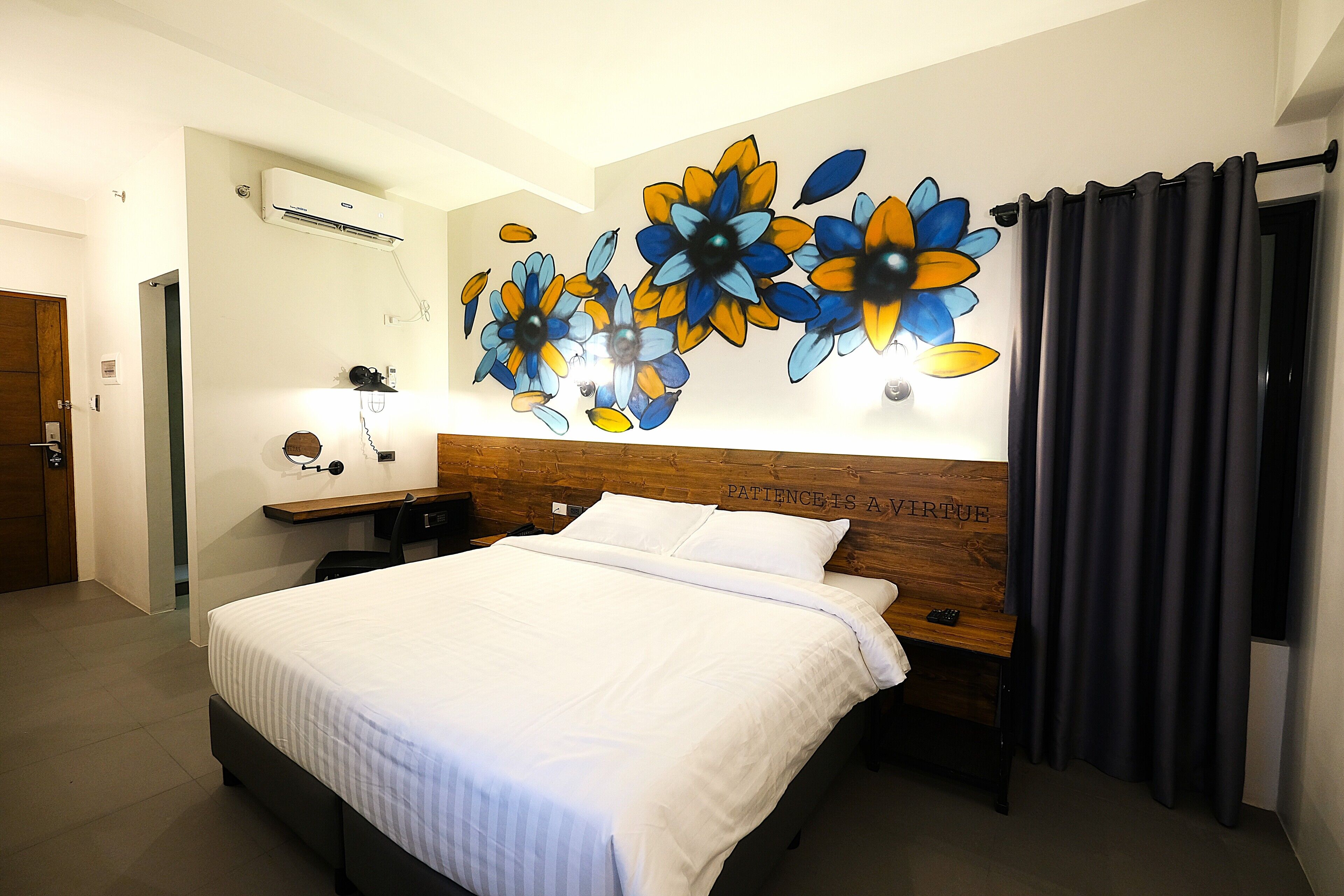 A hotel room with a bed, a desk, a wall-mounted air conditioner, and a decorative floral wall art piece.