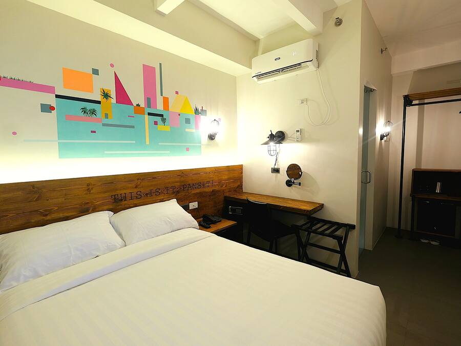 Una habitación de hotel con una cama grande, un escritorio y una silla. Hay un aire acondicionado de pared y una puerta con un zapatero.