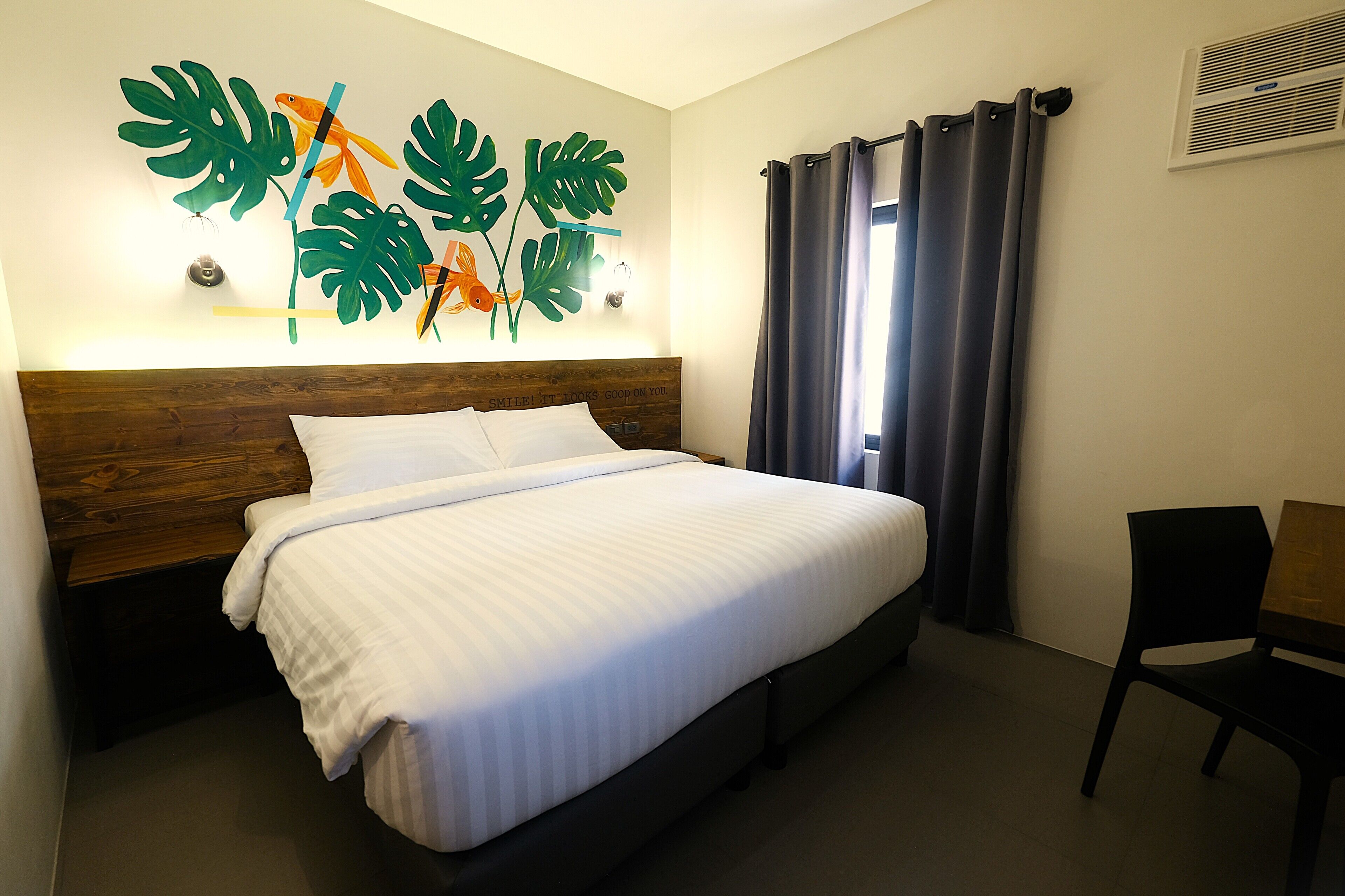 A neatly made bed with a wooden headboard, a wall-mounted artwork of tropical leaves and fish, a window with dark curtains, and a black chair.
