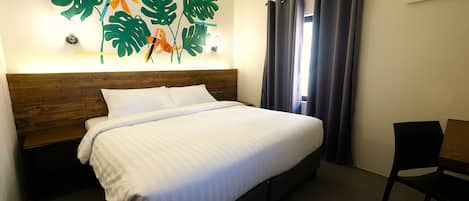 Una cama bien hecha con cabecera de madera, una obra de arte en la pared con hojas tropicales y peces, una ventana con cortinas oscuras y una silla negra.