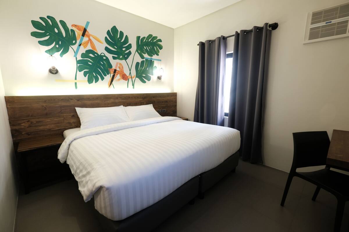 A neatly made bed with a wooden headboard, a wall-mounted artwork of tropical leaves, and a window with curtains.