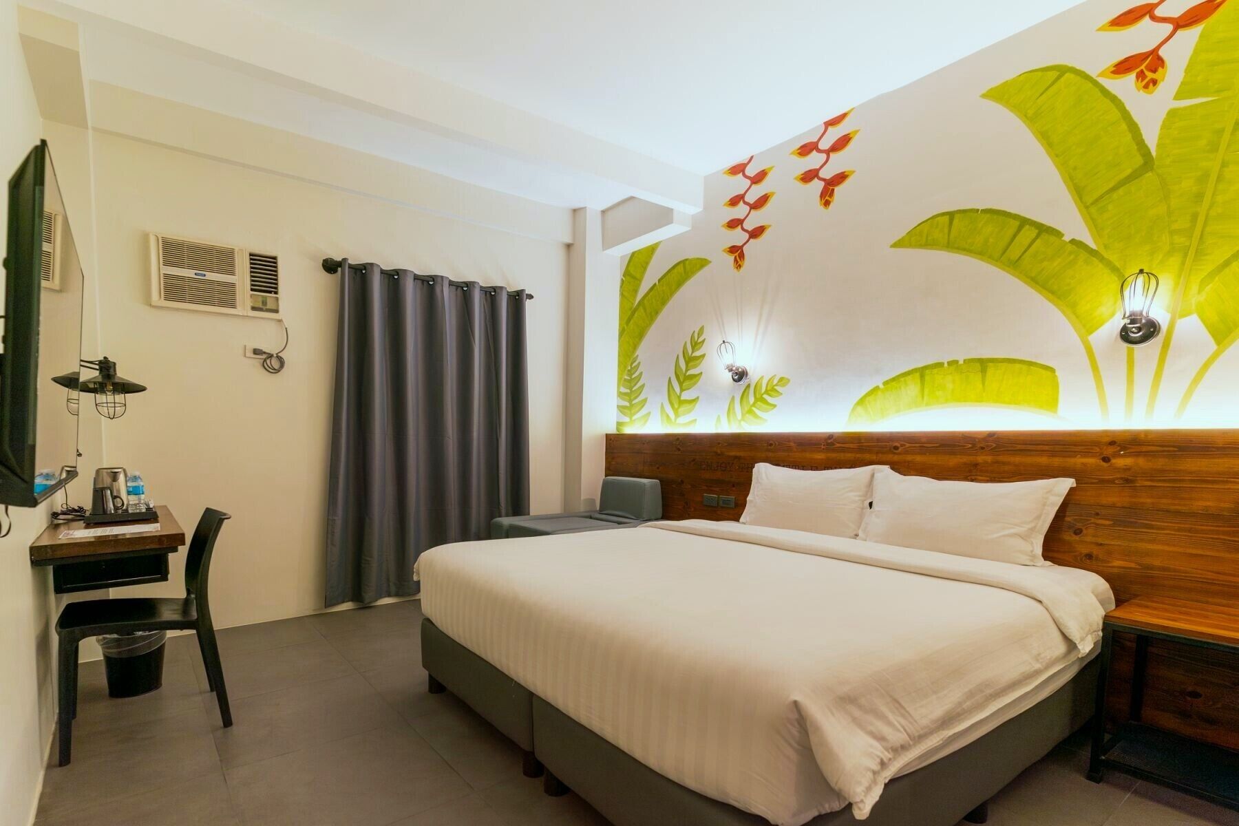 A hotel room with a bed, a desk with a chair, a wall-mounted air conditioner, and a decorative wall with leaf patterns.