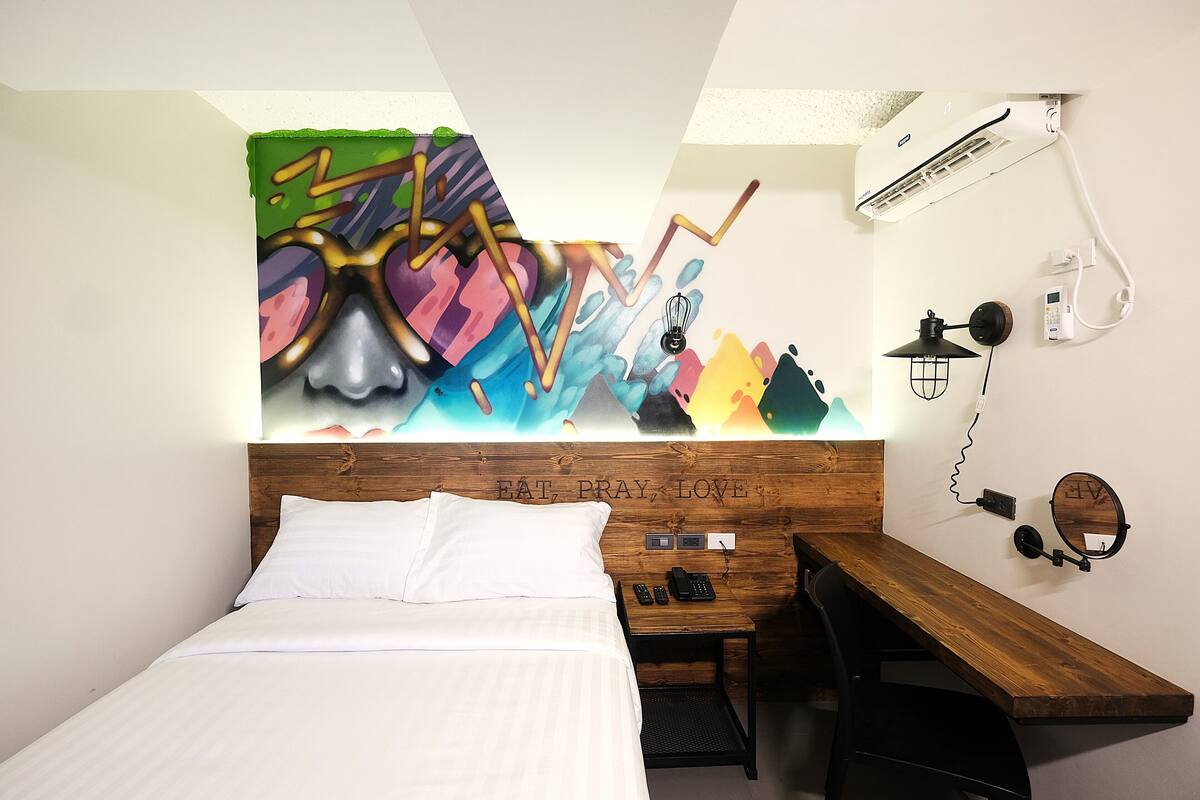 A modern hotel room with a bed, a wooden desk, a wall-mounted phone, and a colorful abstract painting.