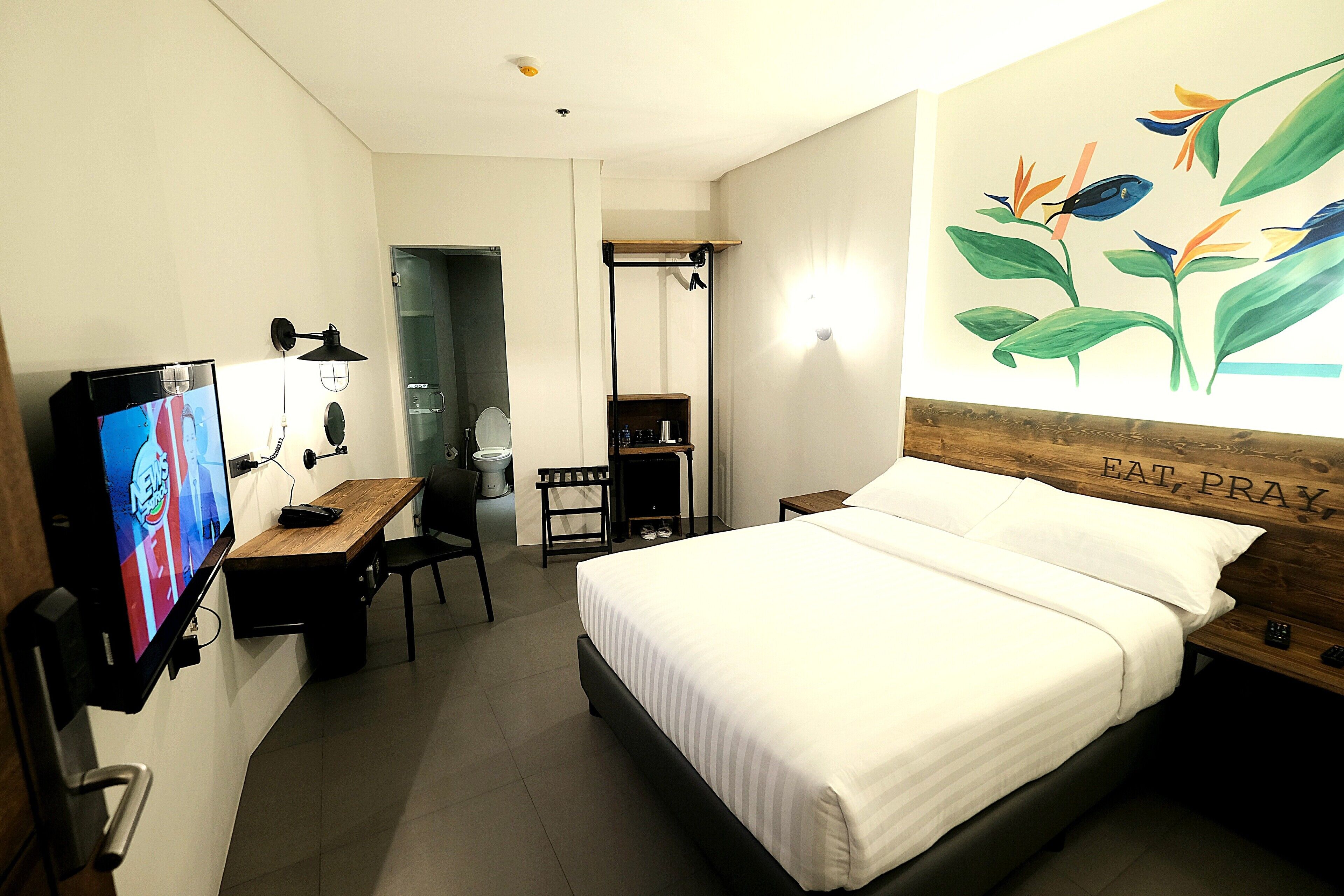 A hotel room with a bed, a desk with a TV, a mirror, and a wall with a plant mural.
