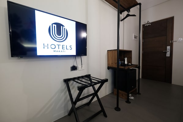 Habitación de hotel con un televisor de pantalla plana mostrando el logotipo de 'HOTELES MAKATI', un perchero de metal negro y un armario de madera con una cafetera.