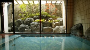 An indoor swimming pool with a tiled floor, surrounded by large rocks and a view of a garden with trees and a waterfall. - Nakaya Ryokan (Minakami)