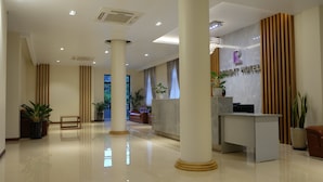 Um saguão de hotel moderno, com uma mesa de recepção, colunas imponentes e plantas em vasos.