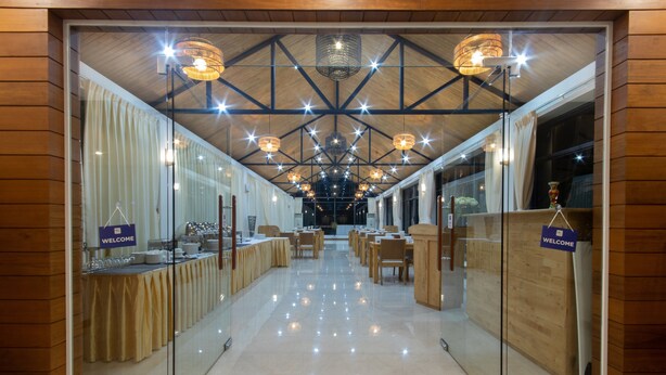 Uma ampla sala de banquetes, bem iluminada, com teto alto e lustres modernos. Mesas estão dispostas para refeições, e há placas indicando a entrada.