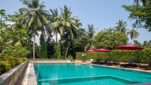 Piscina com espreguiçadeiras e guarda-sóis vermelhos, rodeada por palmeiras e vegetação.