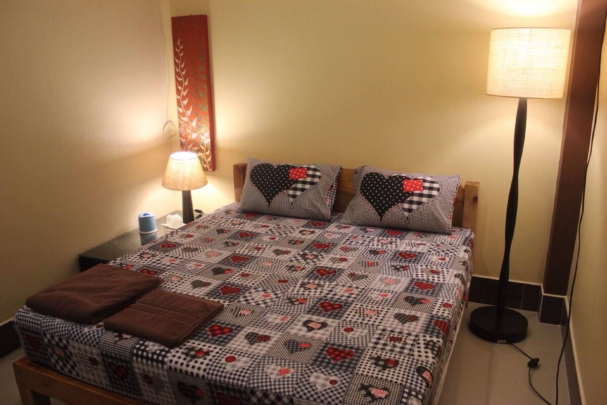 A neatly made bed with a checkered quilt, two heart-patterned pillows, a brown throw blanket, a bedside lamp, and a wall decoration.