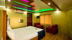 Habitación de hotel con cama, un sillón rojo, una segunda silla roja, televisor de pantalla plana e iluminación LED de colores en el techo.
