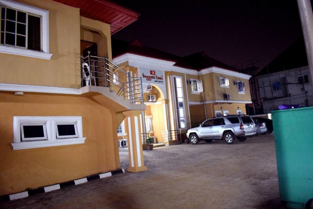 A hotel with a yellow facade, a balcony, and a sign that reads "Burgatti Hotel." There are cars parked in front and a green trash bin to the right.