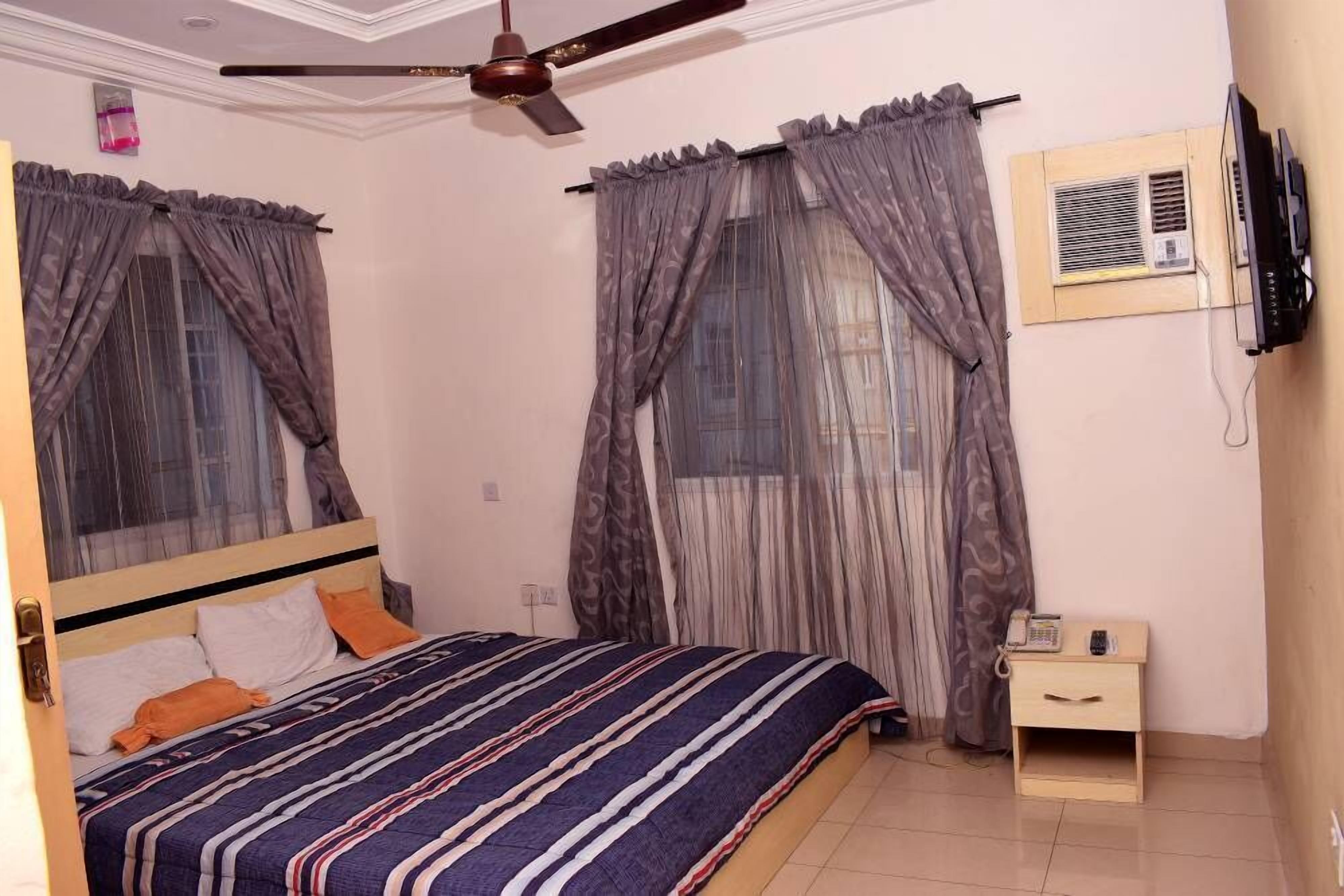 A bedroom with a bed, curtains, a ceiling fan, a bedside table with a phone, and a TV mounted on the wall.