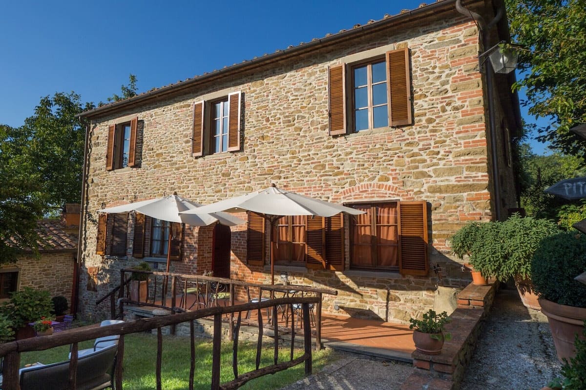 A stone building with wooden shutters, outdoor seating with umbrellas, and a small staircase leading to the entrance.