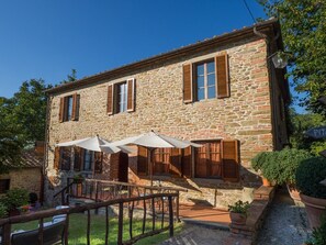 Un edificio de piedra con contraventanas de madera, asientos exteriores con sombrillas y una pequeña escalera que conduce a la entrada.