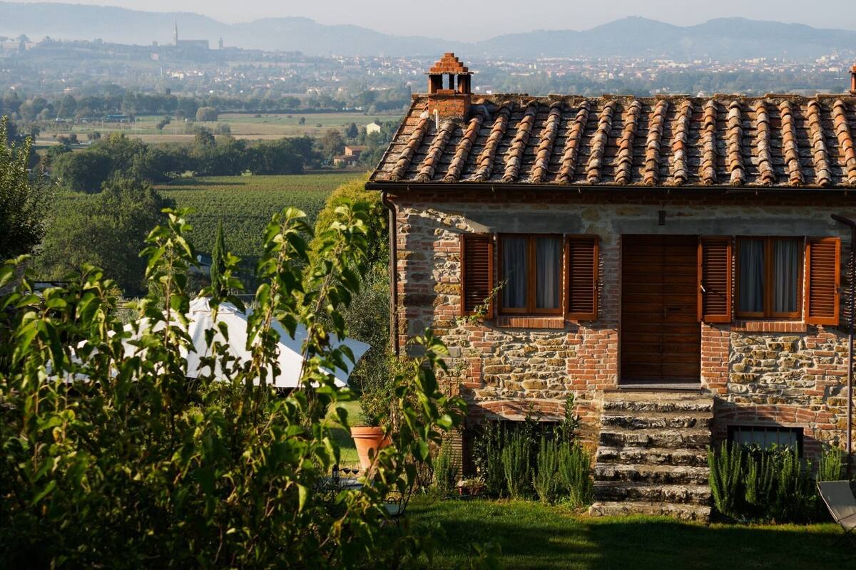 A stone house with a tiled roof, wooden shutters, and a view of a distant town and hills.