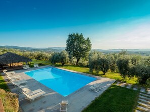 Una piscina rectangular con sillas de descanso y una estructura de techo de paja, rodeada de olivos y vistas a colinas distantes.