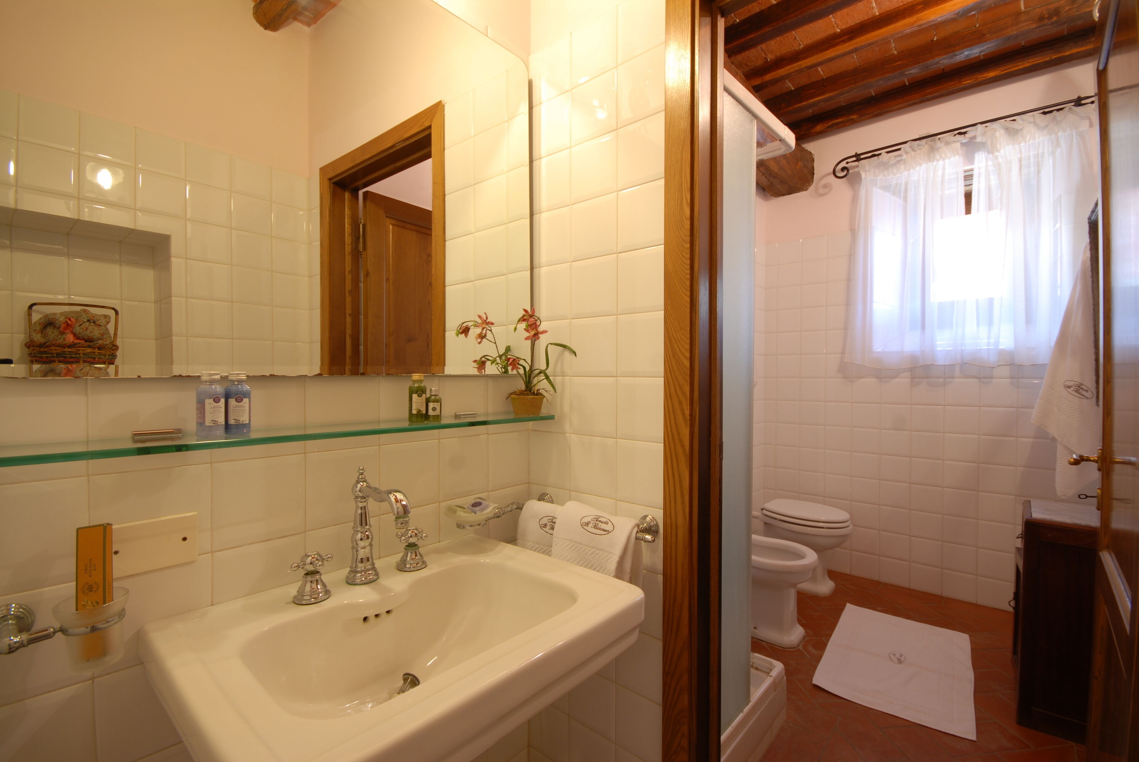 A bathroom with a white sink, mirror, and toilet. There is a glass shelf above the sink with toiletries.
