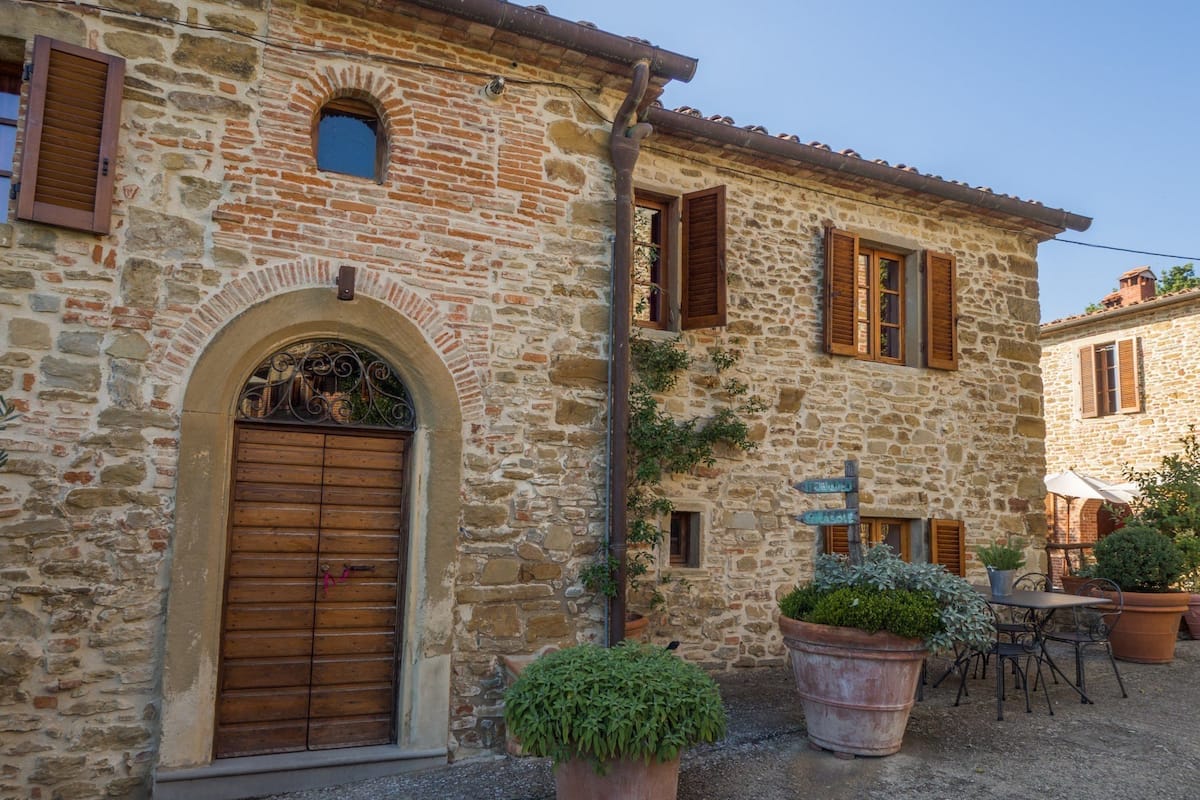 A stone building with a wooden door and windows, outdoor seating area with potted plants, and a signpost with directions.