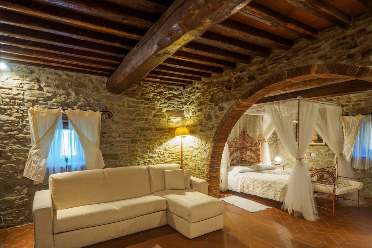 A cozy room with a stone wall, a wooden ceiling, a white sofa, a bed with a canopy, and a floor lamp.
