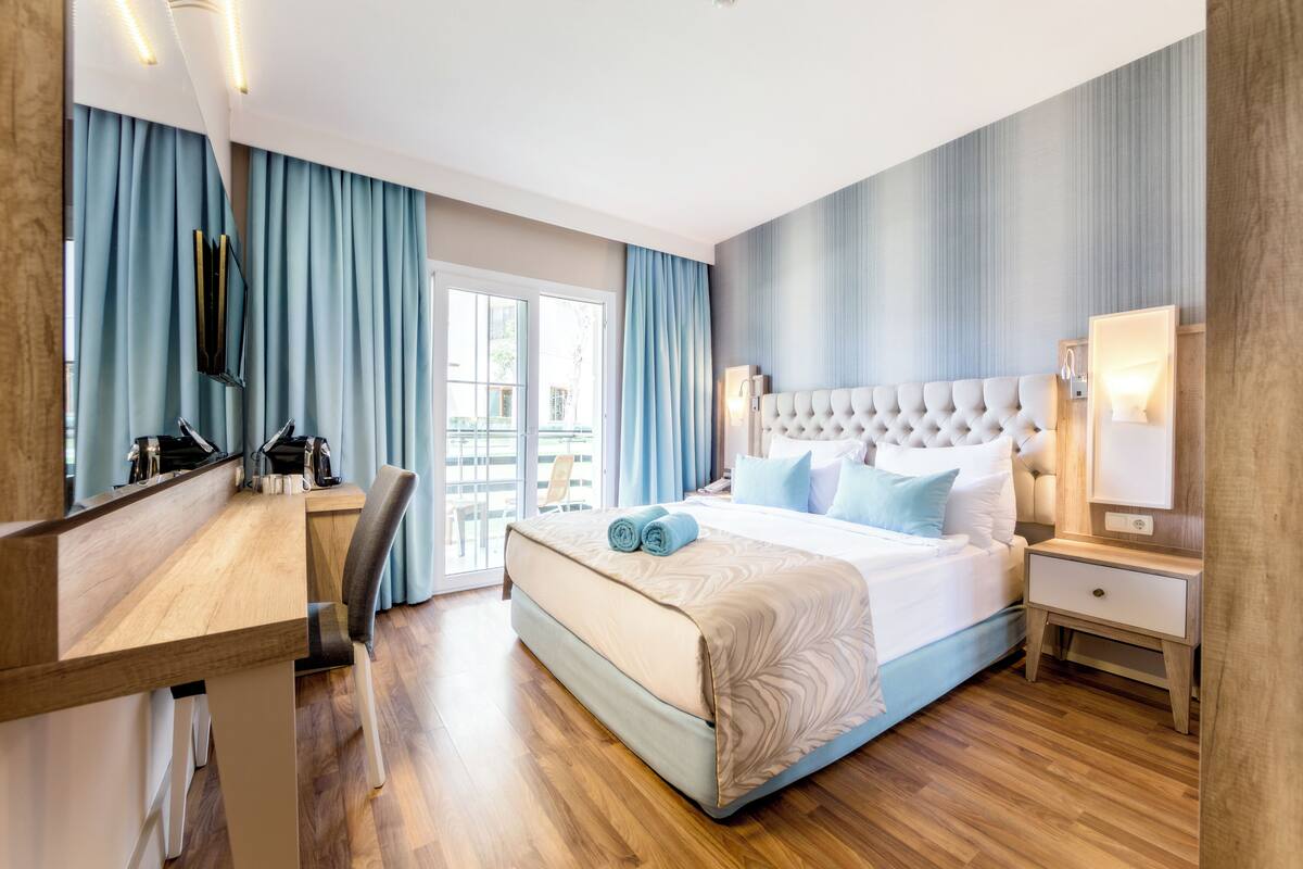 A hotel room with a large bed, a desk, a chair, a TV, and a sliding glass door to the outside.