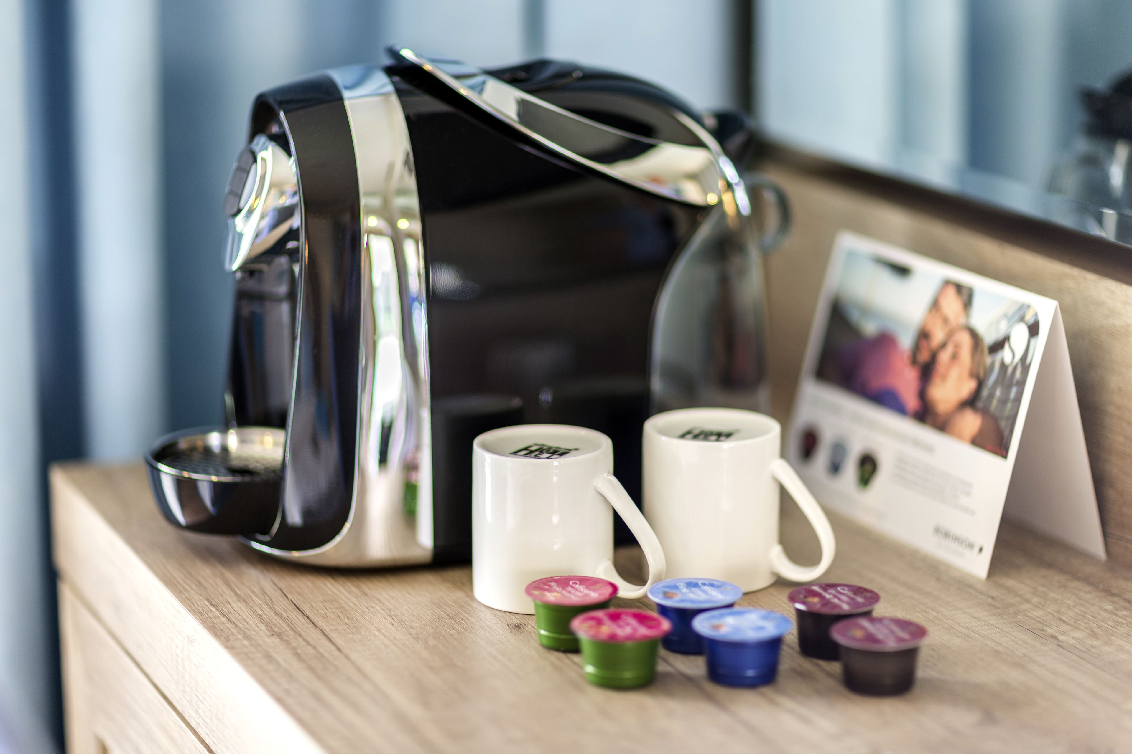 Une machine à café, deux tasses blanches et plusieurs capsules de café colorées sur une surface en bois.