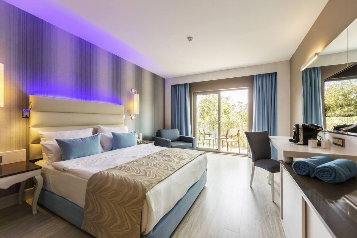 A modern hotel room with a large bed, a desk, and a balcony with a table and chairs.