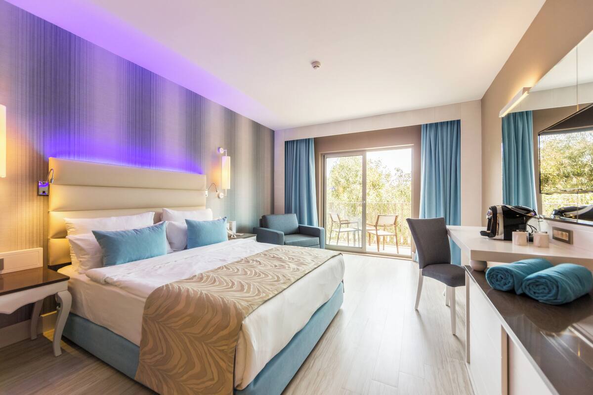 A modern hotel room with a large bed, a desk, and a balcony with a table and chairs.