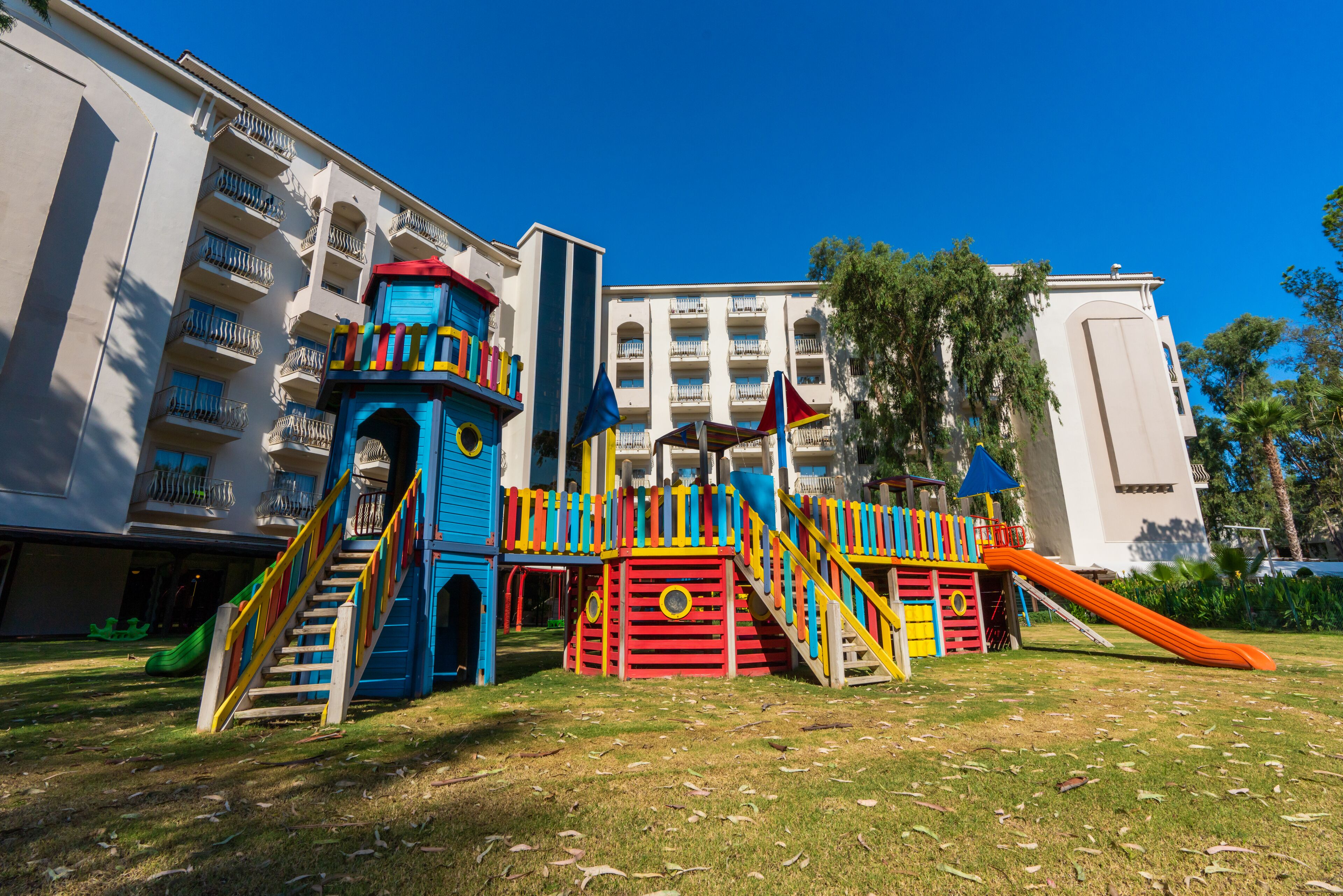 Un parque infantil con estructuras de juego coloridas y un tobogán, rodeado de césped y un edificio con balcones.