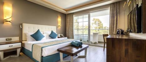 A hotel room with a large bed, bedside tables, a wooden bench, a desk, a TV, and a balcony with a view.