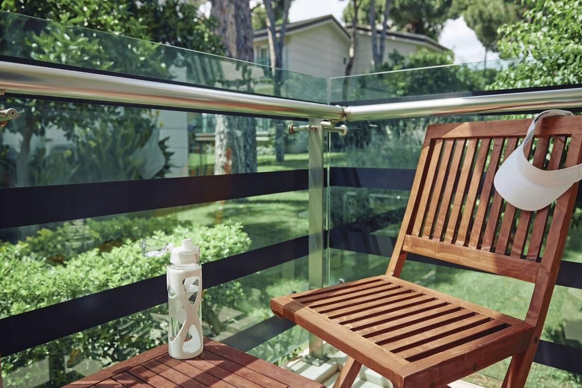 A wooden chair and a white water bottle on a balcony with a glass railing, overlooking a green landscape.