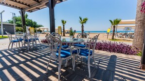 A covered outdoor seating area with white metal chairs and blue cushions, overlooking a beach with palm trees and a clear sky. - TUI MAGIC LIFE Masmavi - All inclusive (Serik)