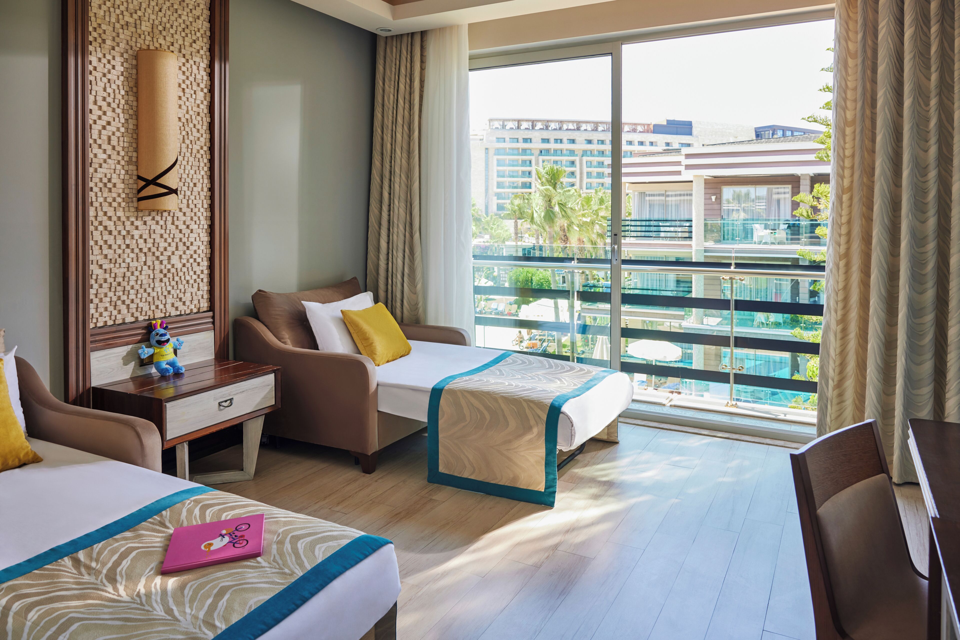 Habitación de hotel con dos camas, un escritorio, una silla y un ventanal con vistas a un edificio y palmeras.