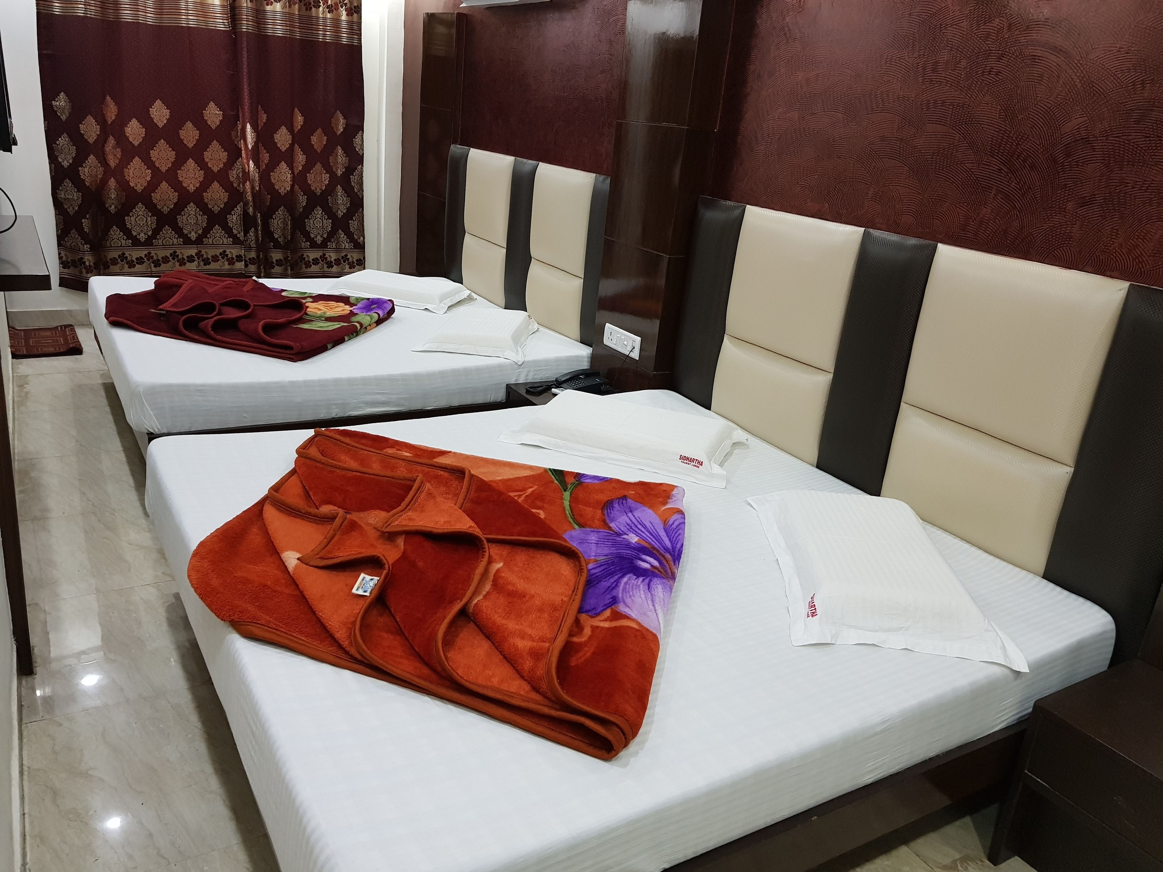A hotel room with two beds, each with a white mattress and a headboard. There is a red and purple floral blanket on one bed and a white sheet on the other.