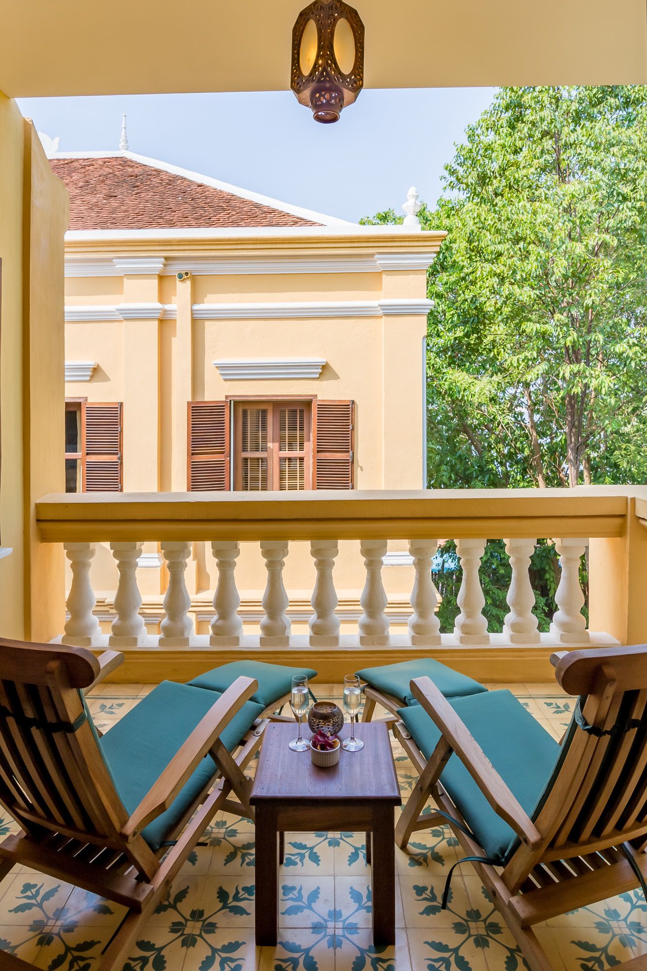 A balcony with wooden chairs and a table, overlooking a building with a tiled roof and a tree.