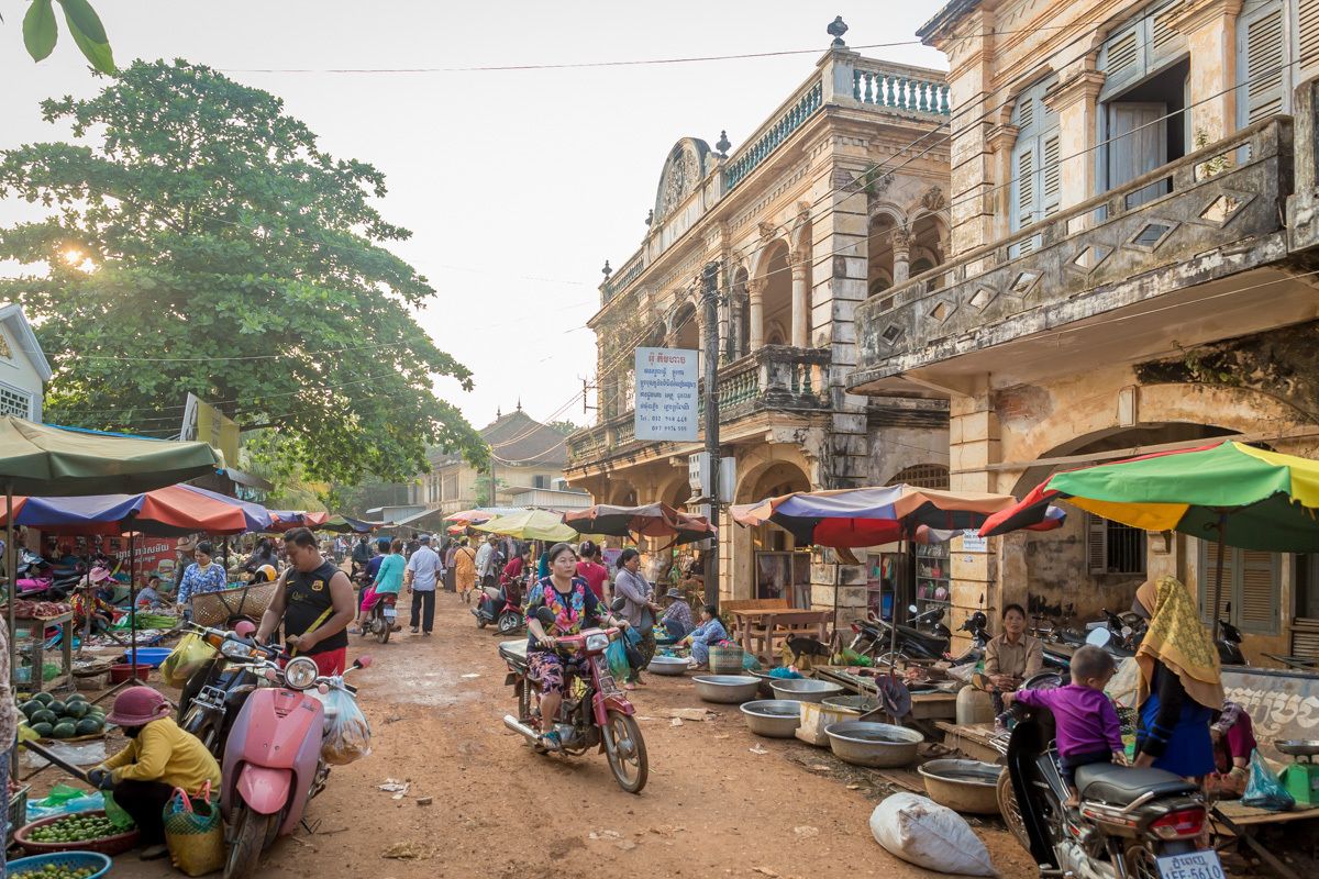 A bustling street market with vendors and shoppers, surrounded by historic buildings with balconies and arches.