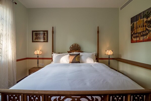 Cama bem arrumada com lençóis brancos, duas luminárias de cabeceira, um quadro na parede e uma pintura que retrata um templo.