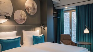 Une chambre d’h ôtel moderne dotée d’un grand lit, d’une tête de lit agrémentée d’œuvres d’art mural, d’un coin salon avec un fauteuil et une table, et offrant une vue sur un bâtiment en briques par la fenêtre.