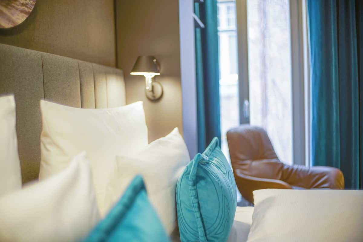 A hotel room with a bed, pillows, a lamp, and a view of the outdoors through a window.