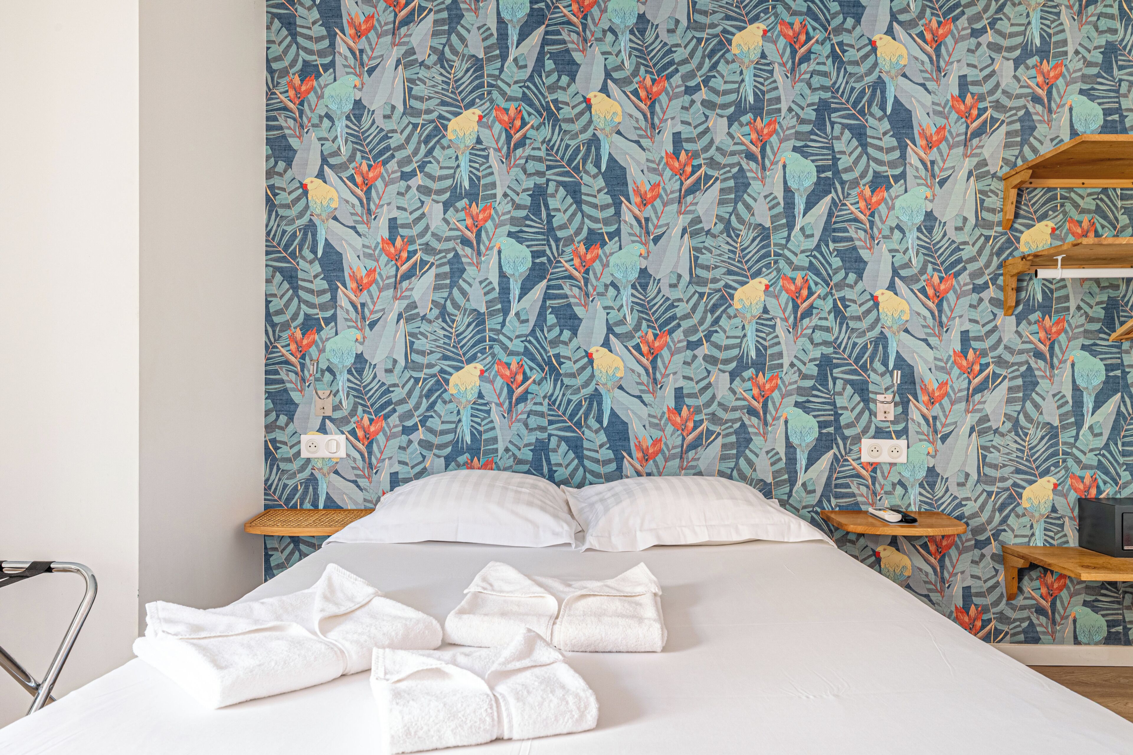 Une chambre d’hôtel avec un grand lit, deux tables de chevet, une étagère en bois et un mur recouvert d’un papier peint représentant des feuilles et des oiseaux tropicaux.