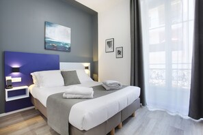 Una habitación de hotel moderna con una cama grande, una mesita de noche con lámpara, un cuadro en la pared y una ventana con vistas.