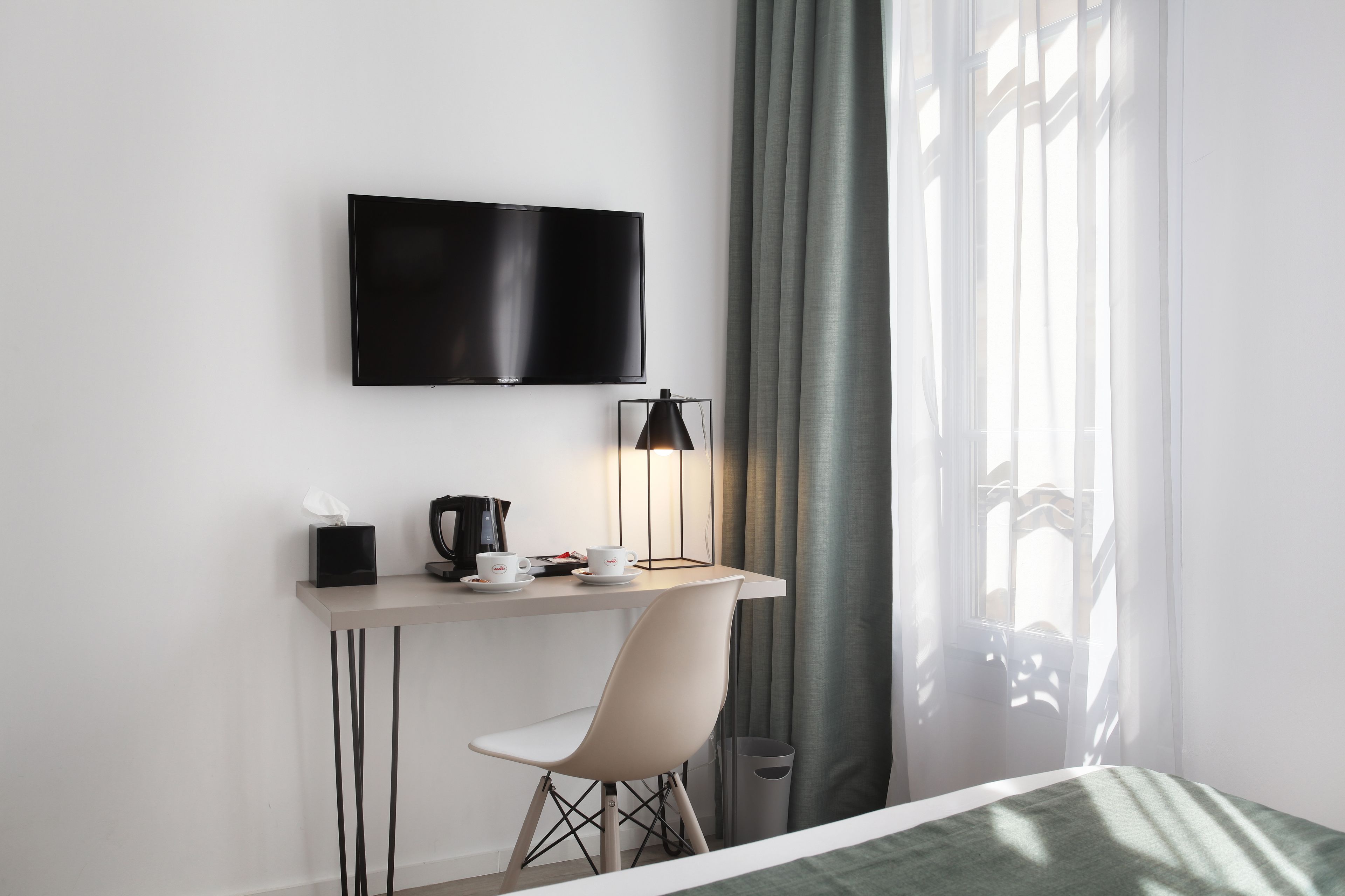 A modern hotel room with a flat-screen TV mounted on the wall, a desk with a lamp, a chair, and a bed with a green bedspread.