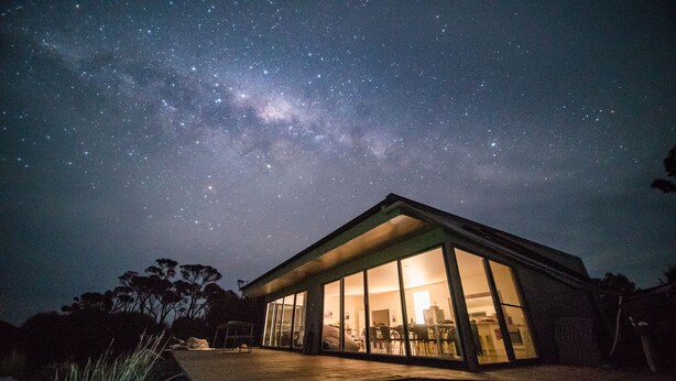 Une maison moderne dotée de grandes baies vitrées, d’une terrasse et offrant une vue sur le ciel nocturne.
