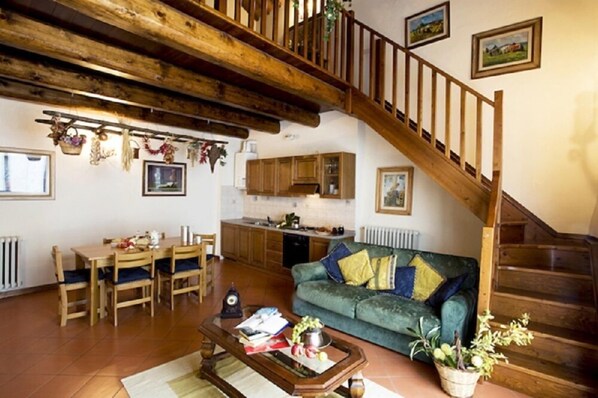 A spacious living area with a wooden staircase, a green sofa, a dining table with chairs, and a kitchen in the background. - Agriturismo Petrognano (Reggello)