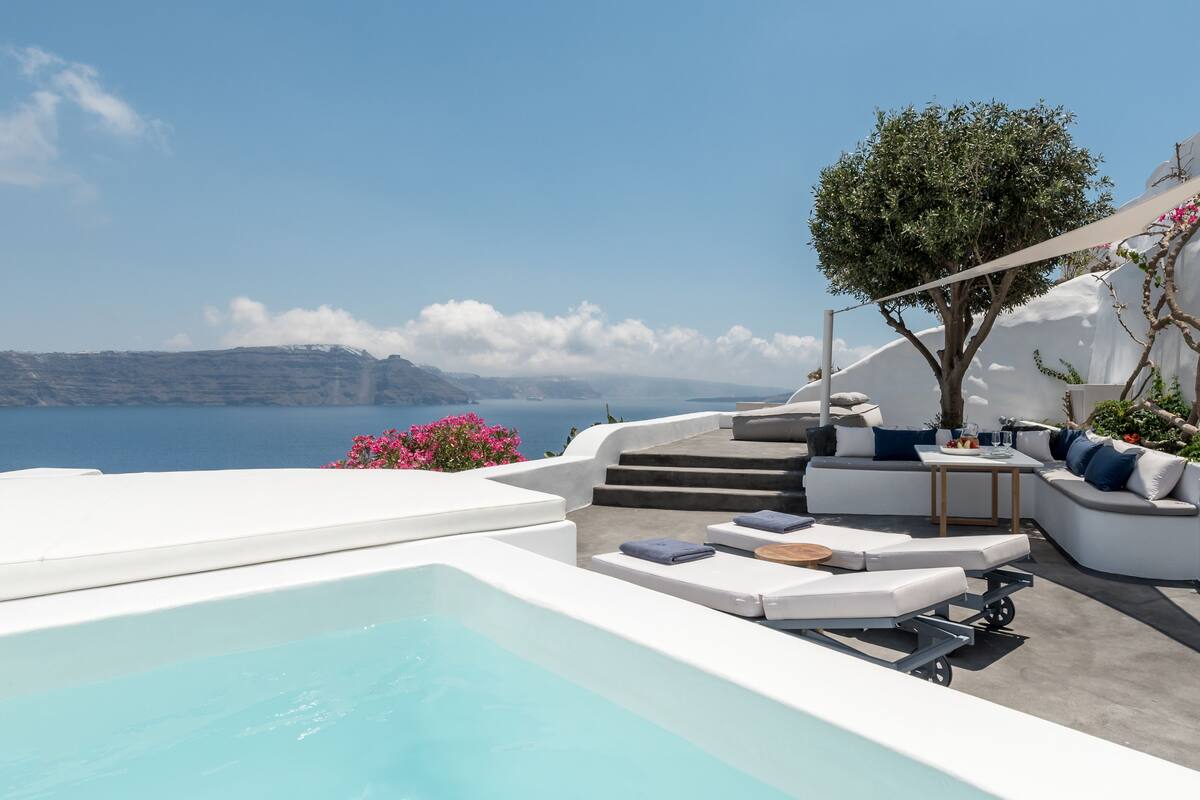 Un área de piscina moderna al aire libre con vista al mar, mobiliario de sala de estar blanco y un árbol.