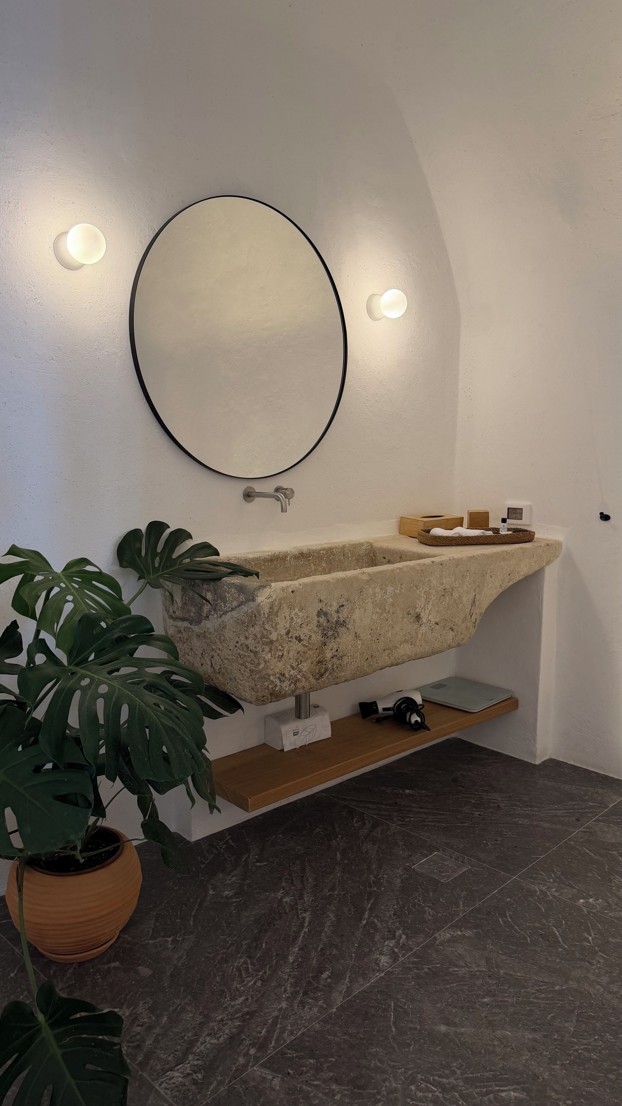 Un baño con lavamanos de piedra, un espejo redondo, una planta en maceta y un pequeño estante con artículos de aseo.