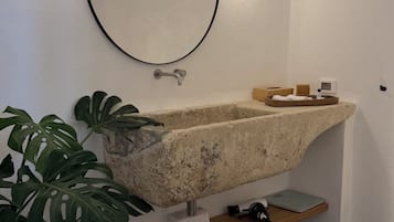 Un baño con lavamanos de piedra, un espejo redondo, una planta en maceta y un pequeño estante con artículos de aseo.