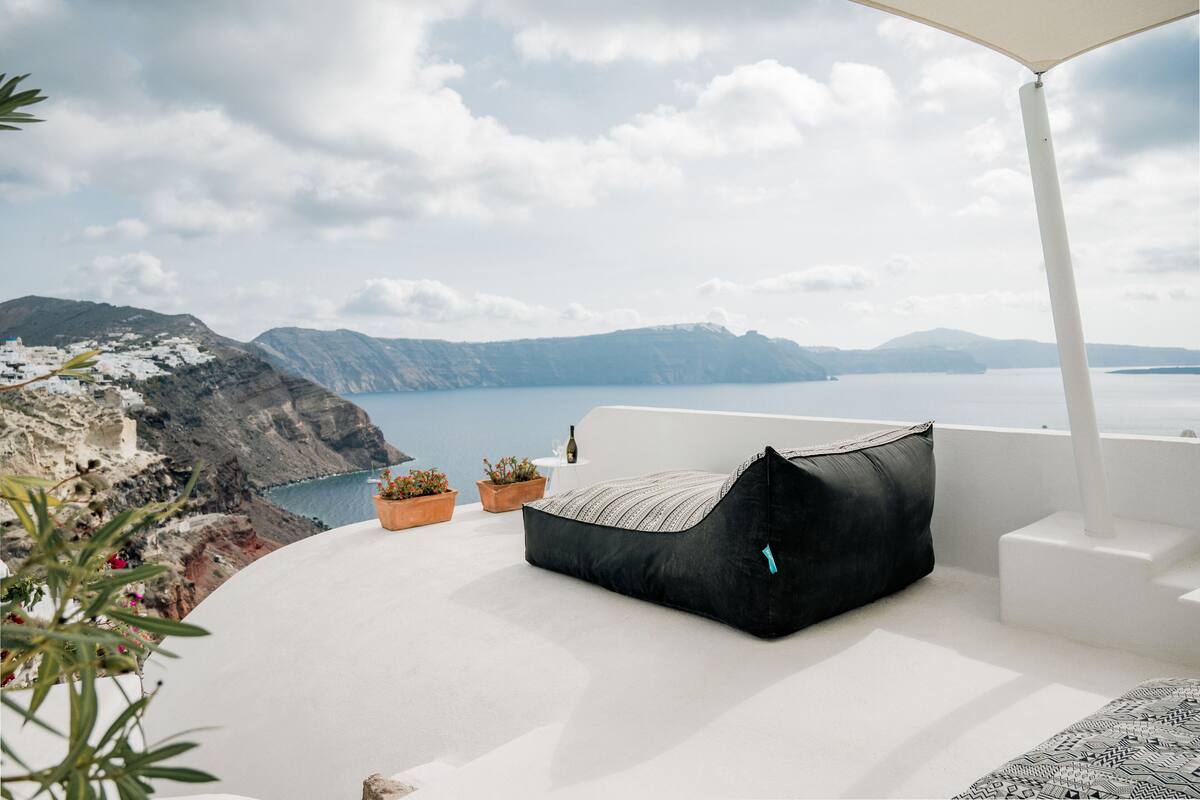 Un espacio exterior con asientos blancos, un sillón de mimbre negro, plantas en macetas y vistas al mar y las montañas.