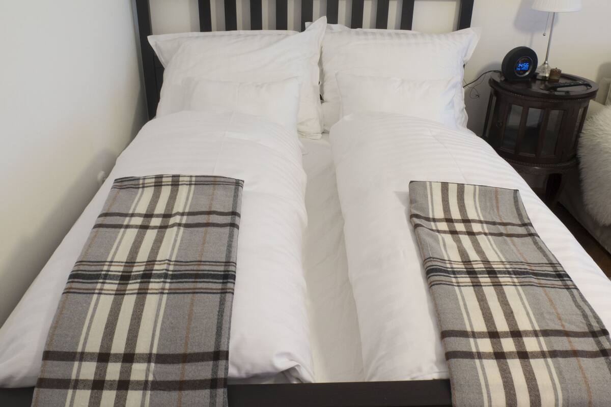 A bed with white bedding, a plaid blanket, and a bedside table with a lamp and a clock.
