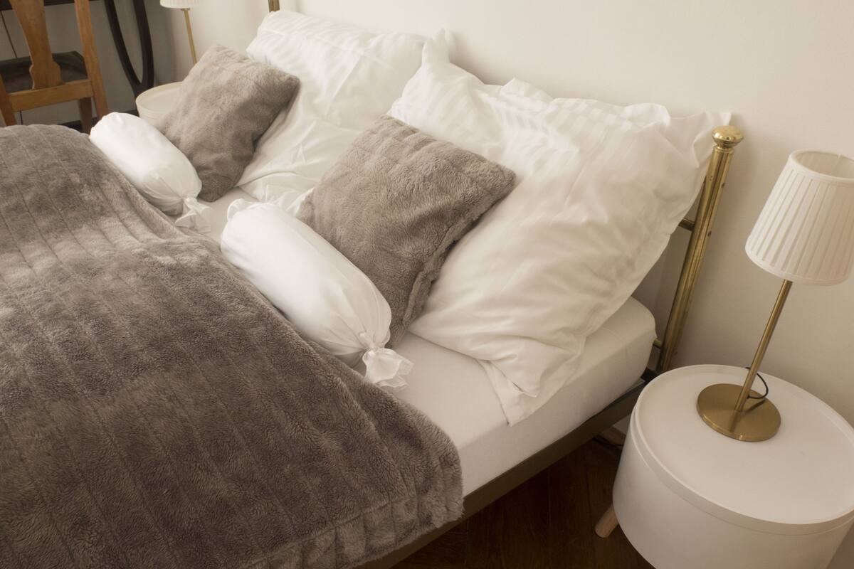 A neatly made bed with a grey blanket and white pillows, a bedside lamp, and a small white nightstand.